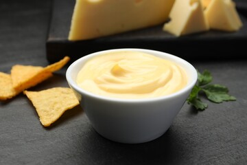 Tasty dipping cheese sauce in bowl, nacho chips and parsley on black table, closeup