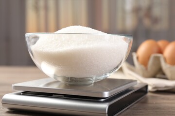 Digital kitchen scale with bowl of sugar on wooden table indoors, closeup