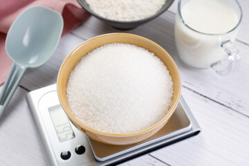 Digital kitchen scale with bowl of sugar and other products on white wooden table, closeup