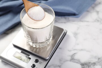 Putting sugar into glass on digital kitchen scale at white marble table, closeup. Space for text