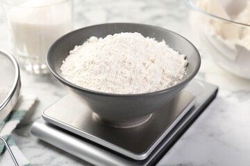 Digital kitchen scale with bowl of flour and other products on white marble table, closeup
