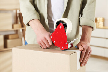 Man using dispenser with adhesive tape to seal cardboard box indoors, closeup