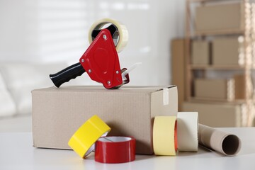Dispenser with adhesive tape rolls and cardboard box on desk indoors