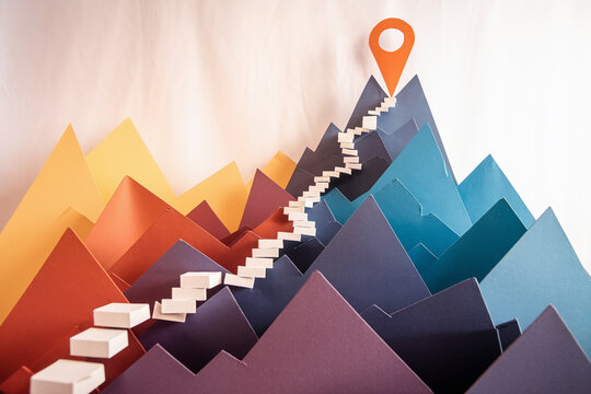 Abstract paper mountainscape with steps leading to a peak marked by a location pin, symbolizing achievement and progress, ideal for goal-oriented concepts