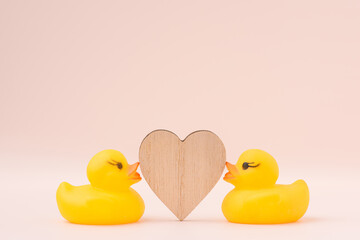 Two yellow rubber ducks facing a wooden heart on soft pink background. Studio flat lay composition and gentle light shadow.