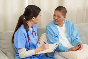 Obraz premium Asian female physiotherapist wearing blue uniform holding pen and clipboard talking with senior asian female patient on sofa during home care session discussing treatment recovery medical assistance