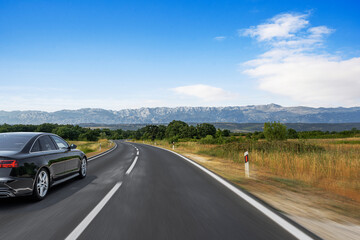 A sleek black car speeds along a curving highway through the countryside, with majestic mountains rising beneath a vibrant blue sky.