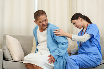Fototapeta premium Young asian female physical therapist assisting elderly female patient with back pain during rehabilitation session on sofa applying hands-on care promoting support recovery posture improvement