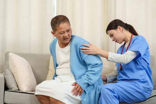 Young asian female physical therapist assisting elderly female patient with back pain during rehabilitation session on sofa applying hands-on care promoting support recovery posture improvement