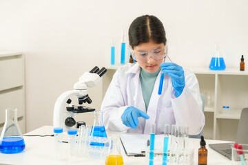 Teenage caucasian schoolgirl in science class wearing lab coat gloves goggles holding test tube with blue liquid while pointing at data on clipboard surrounded by laboratory tools and glassware