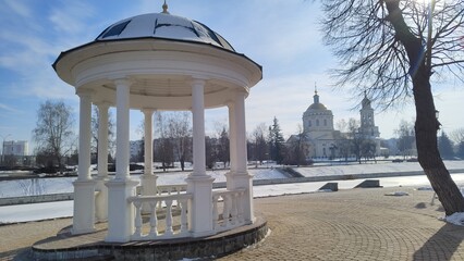 Rotunda on left bank of Orlik River with beautiful view of Cathedral of Archangel Michael in Orel city. Old white gazebo on background of river and Orthodox church on sunny winter day