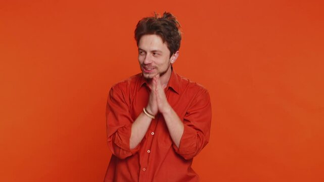 Sneaky cunning Caucasian young man with tricky face gesticulating and scheming evil plan, thinking over devious villain idea, cheats, jokes, pranks. Handsome guy isolated on orange studio background