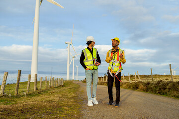Two engineers walking and discussing next to wind turbines in a wind farm, wearing safety vests and...