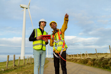 Two engineers wearing safety vests and helmets are inspecting wind turbines, one holding a digital tablet and the other pointing towards a turbine