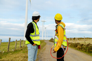 Two engineers wearing safety vests and helmets are discussing next to wind turbines, showcasing...