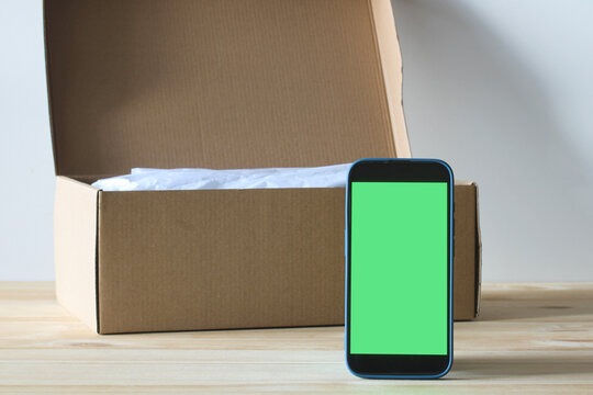 Smartphone with green screen standing next to open cardboard box on wooden table - Powered by Adobe
