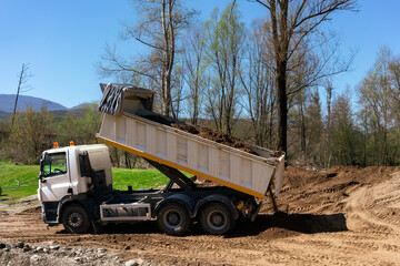 White dump truck unloading soil at a construction site. Heavy machinery performing earth works, backfilling for a landscape architecture project. Industrial earthmoving operation. © Christos