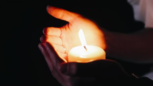 The woman covers the flame from the candle with her hands, not letting it go out. The candle flame illuminates the woman's hands. Close-up shooting