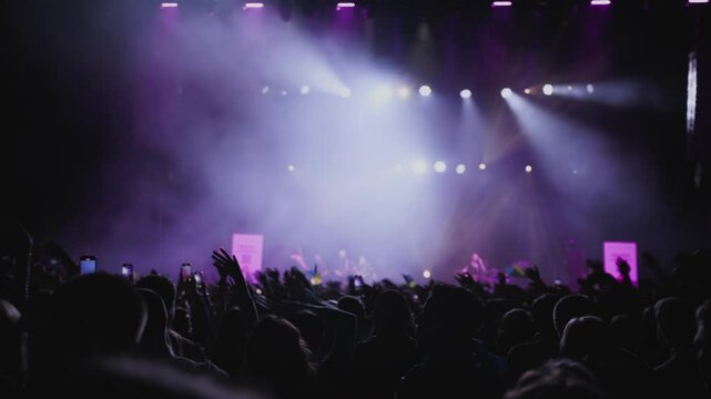 Perfect light show. Fans with rased your hands and wave for music against background white, blue and magenta spotlight on stage. Medium shot.