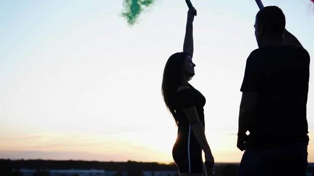 In the evening, at sunset, two lovers are having fun with smoke bombs on the roof.
