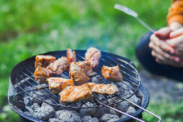 Grilled meat pieces sizzling on a barbecue grate over hot coals, with people nearby preparing skewers, set against a green natural outdoor background.
