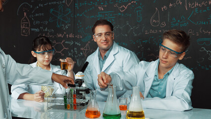 Creative teacher pointing chemistry at blackboard while talking to highschool boy at table with microscope and test tube with colored liquid. Young student wearing lab coat in STEM class. Edification.
