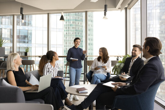 Teambuilding activity. Young Indian girl business speaker leader trainer gather multiethnic coworkers group around office table on meeting seminar having fun on workshop briefing friendly negotiations