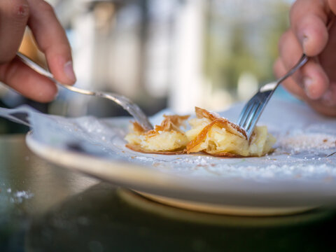 Closeup of hands eating traditional Greek bougatsa, a sweet cream pie pastry with filo dough, dusted with powdered sugar and cinnamon. Popular breakfast or snack from Thessaloniki and Serres, Greece.