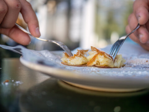 Closeup of hands eating traditional Greek bougatsa, a sweet cream pie pastry with filo dough, dusted with powdered sugar and cinnamon. Popular breakfast or snack from Thessaloniki and Serres, Greece.