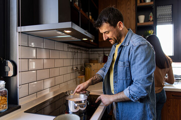 Man stirring food in saucepan on induction stove in modern kitchen, woman washing dishes in...