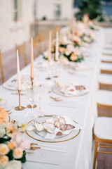 Invitation lies near a napkin on a plate on a set table with bouquets of flowers