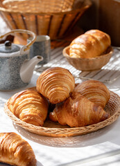 Homemade French butter croissant in wooden basket with cup and pot of tea, cozy ambiance of sunlight of the morning from window shade.