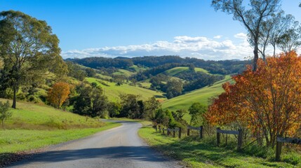A scenic view of a picturesque road winding through lush green hills, with vibrant autumn foliage lining the sides and a clear blue sky above.