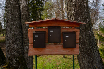 Wooden Stand with Outdoor Mailboxes in Nature. Three dark metal mailboxes mounted on a handmade wooden stand in a natural forested area with spring greenery and trees.