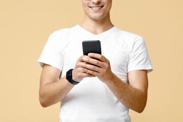 Surfing on smartphone or write in online blog. Attractive smart smiling young man in white t-shirt typing at phone and reading message, isolated on beige background, studio shot, free space