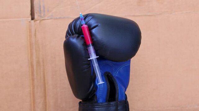Orbit shot of black boxing glove that holds syringe and needle filled with red liquid on cardboard background