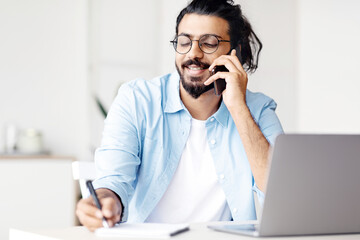 Young Arab Male Entrepreneur Talking On Cellphone And Taking Notes In Office, Handsome Eastern Manager Guy Sitting At Desk With Laptop And Noting Information To Notepad, Speaking With Client