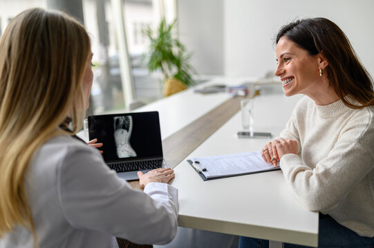Female doctor explaining digital X-ray over laptop to smiling patient during consultation in medical clinic