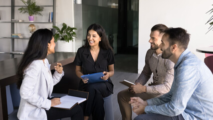Group of young multiethnic teammates talking seated in workspace, laughing, feel satisfied with teamwork, share creative ideas or solutions, take part in briefing in office. Collaborative interaction
