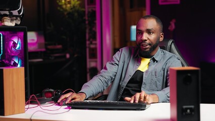 Man in neon lit apartment playing intense videogames on gaming PC at computer desk. Gamer competing...
