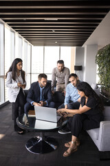 Group of five professionals gathered around laptop in modern office lounge area, collaborating,...
