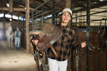 Asian woman rancher carrying horse tack and saddle in horse farm.