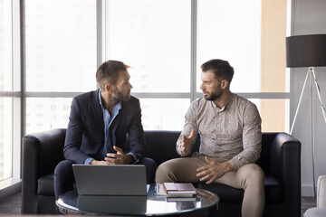 Two serious men, teammates solve work-related matters seated on sofa in company lobby with laptop,...