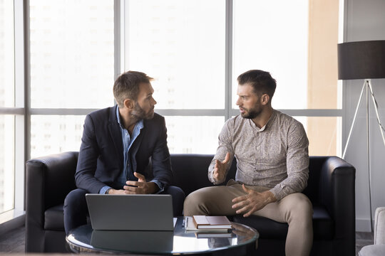 Two serious men, teammates solve work-related matters seated on sofa in company lobby with laptop, collaborating on important decision, engaged in strategic discussion or consultation formal meeting