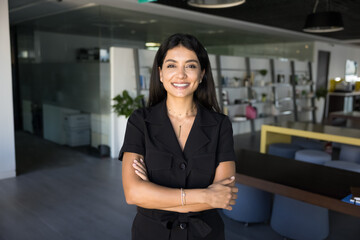 Attractive millennial 35s businesswoman posing in office with her arms-crossed, looks at camera,...