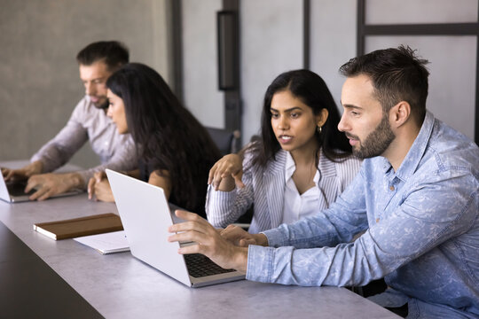 Two multiethnic teammates sit at desk using laptop, busy in strategy or proposal development, drafting strategies, discussing reviewed data, brainstorming, or exploring creative ideas for new project