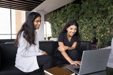 Two businesswomen engaged in lively and friendly interaction in office lounge, laughing, sharing or...
