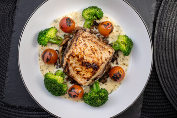 Grilled fish fillet is served with steamed broccoli, grilled cherry tomatoes, creamy sauce and wild rice on a white plate placed on a dark table with a black woven placemat