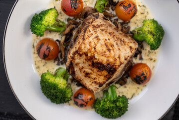 Grilled white fish fillet seasoned and cooked, served with creamy sauce, broccoli and roasted tomatoes, healthy and delicious seafood meal, top view