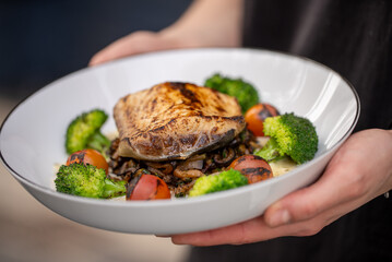 Chef holding a sleek white plate, presenting a vibrant dish of grilled fish accompanied by tender broccoli, juicy cherry tomatoes, and earthy mushrooms, embodying a nutritious and appetizing meal